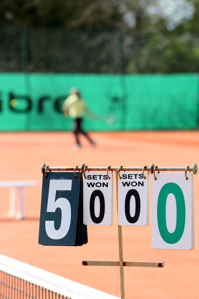 gallery-6 Close-up of tennis scoreboard with blurred player in action on clay court.