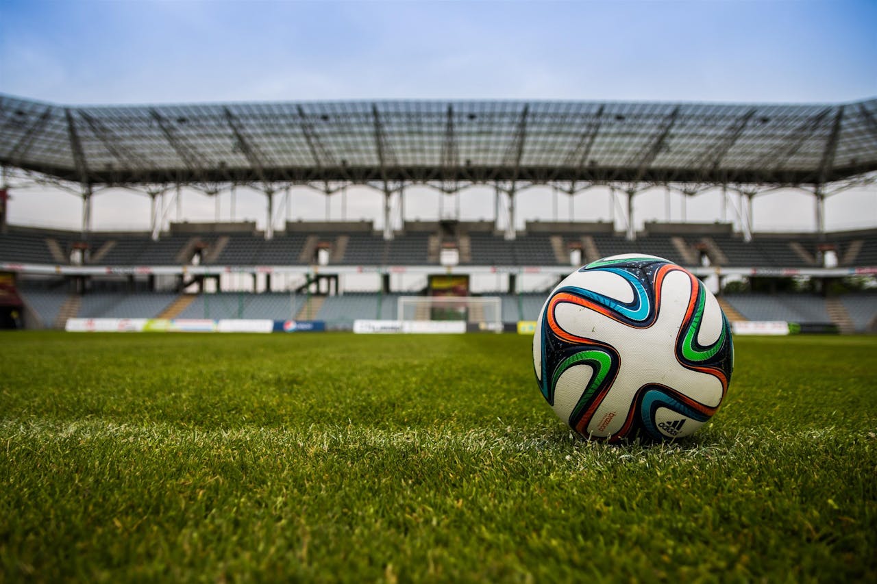 hero-img-02 Close-up of a soccer ball on a lush grass field with an empty stadium in the background.
